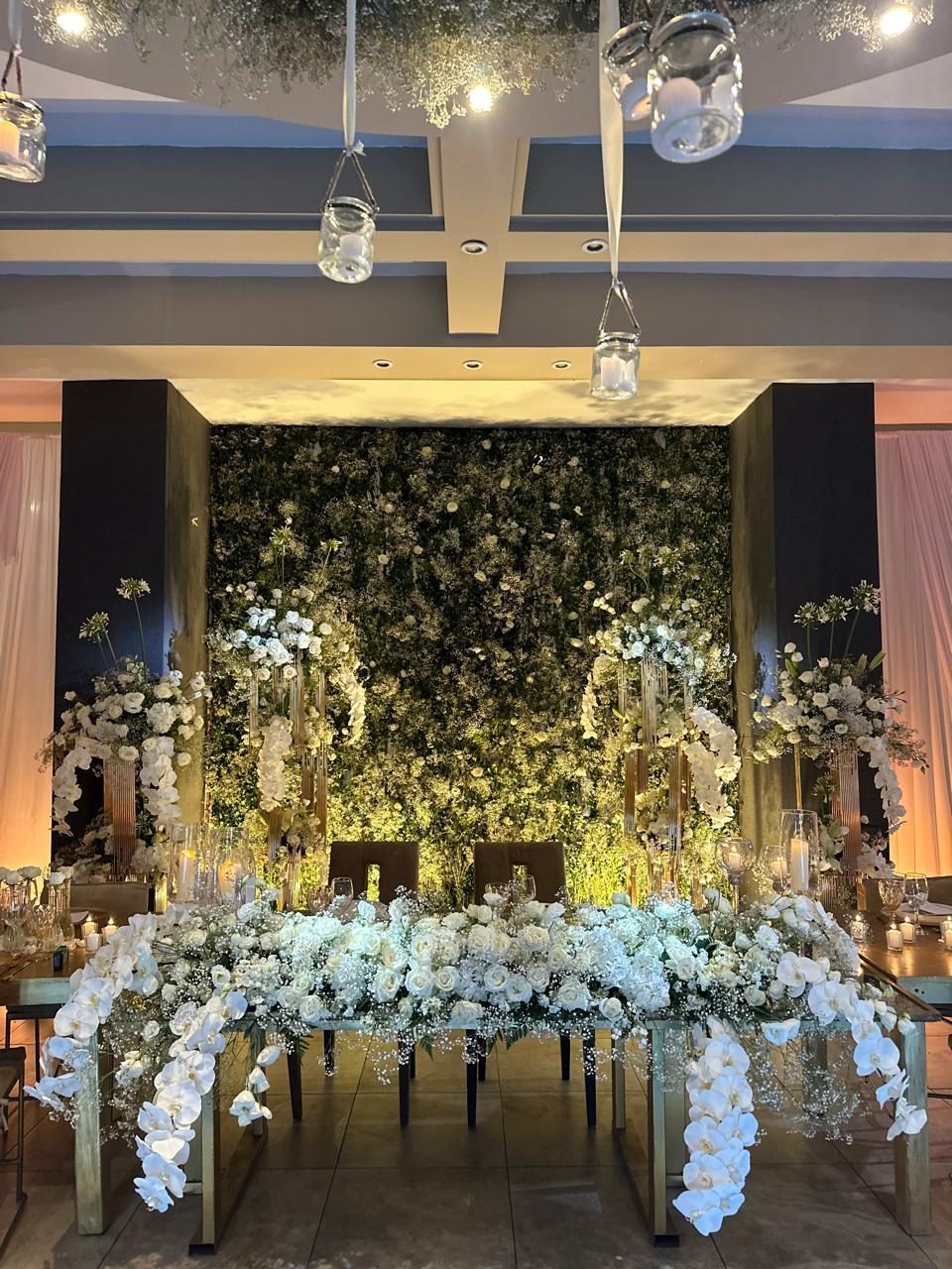 Sweetheart table set against a greenery wall with cascading white orchids and hanging candle vessels