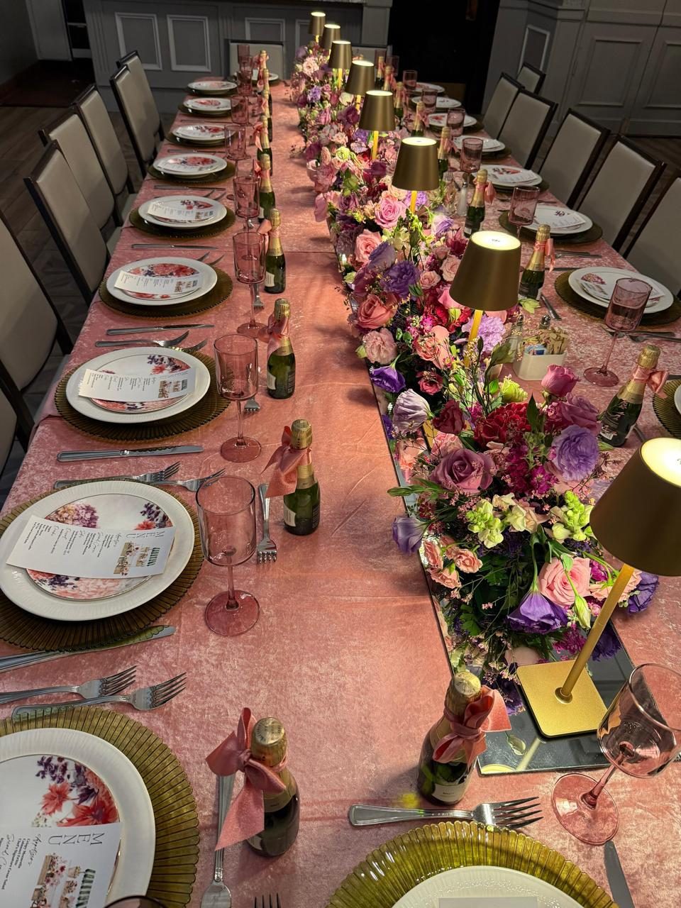 Long reception table with a blush runner, low florals in plum and rose, and gold lamps