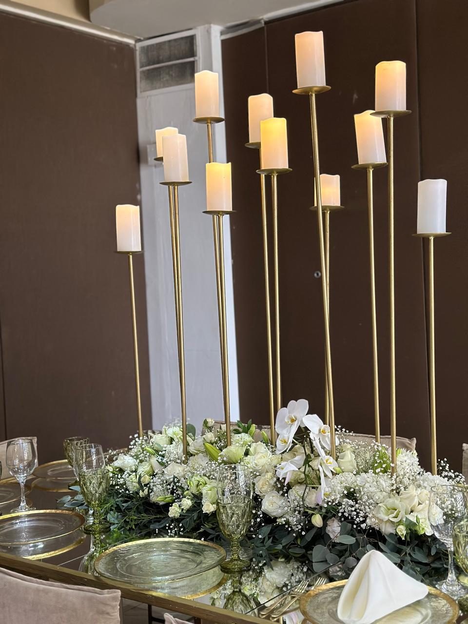 Tall gold candlesticks with white pillar candles above a low arrangement of white orchids and roses
